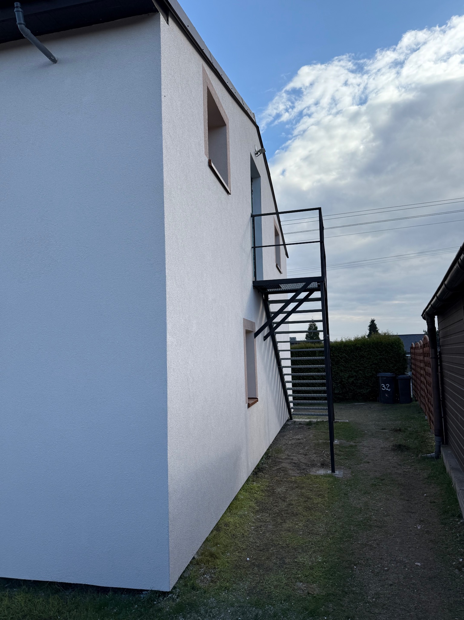 Boczny widok na świeżo otynkowany, dwukondygnacyjny budynek z zewnętrzną, metalową klatką schodową. Biała elewacja kontrastuje z ciemną konstrukcją schodów.