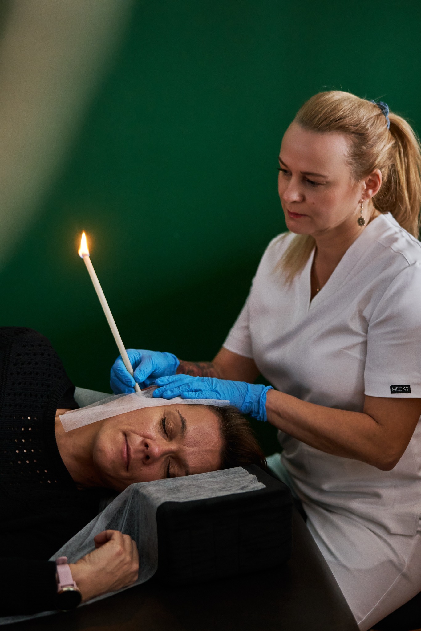 Kobieta poddaje się zabiegowi świecowania uszu przez terapeutkę w niebieskich rękawiczkach. Płomień świecy skierowany ku górze, zielone tło.