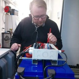 PIOKAMI Rajmund Januszewski - Elektryk w okularach sprawdza instalację w białej skrzynce na dwóch niebieskich akumulatorach, używając czerwonego miernika. Widoczne kable i elementy elektryczne.