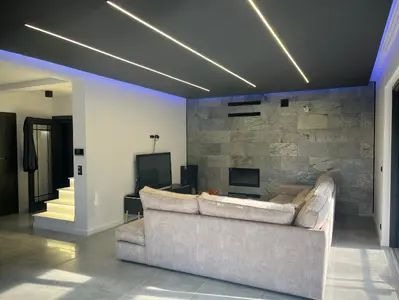 Nowoczesny salon w Kowarach z oświetleniem LED na suficie i podświetleniem. Szara ściana z kamienia, beżowa sofa. Minimalistyczny design.