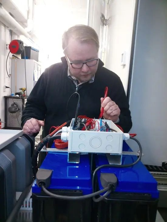 Elektryk w okularach sprawdza instalację w białej skrzynce na dwóch niebieskich akumulatorach, używając czerwonego miernika. Widoczne kable i elementy elektryczne.