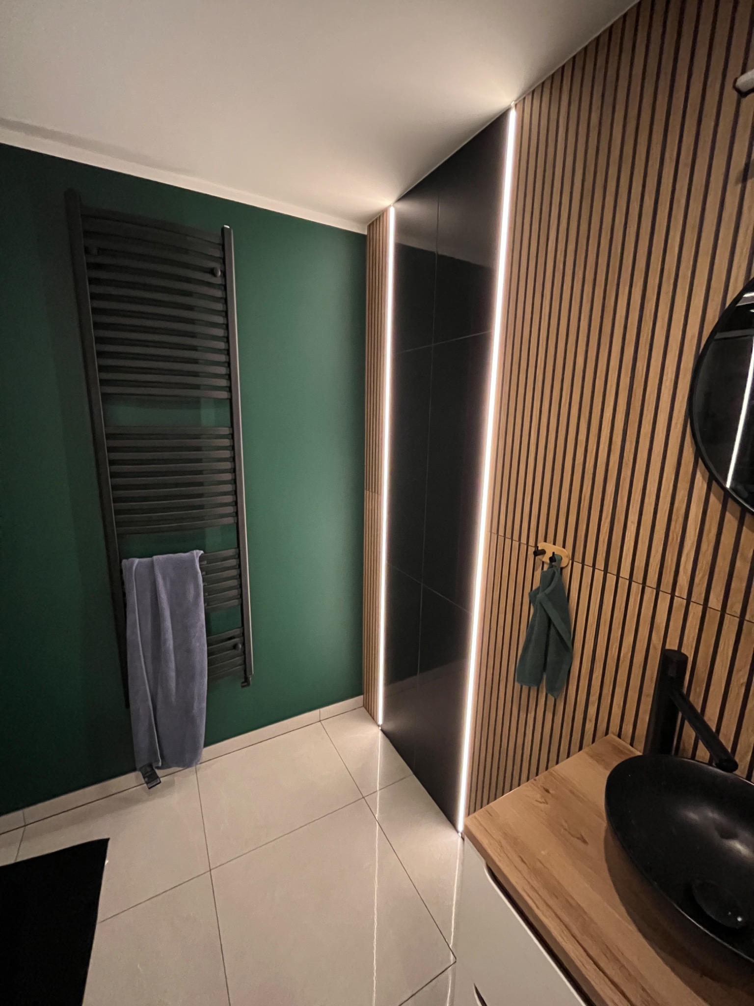 Nowoczesna łazienka z drewnianą ścianą z lameli, podświetleniem LED, czarną umywalką nablatową i grzejnikiem drabinkowym na zielonej ścianie.