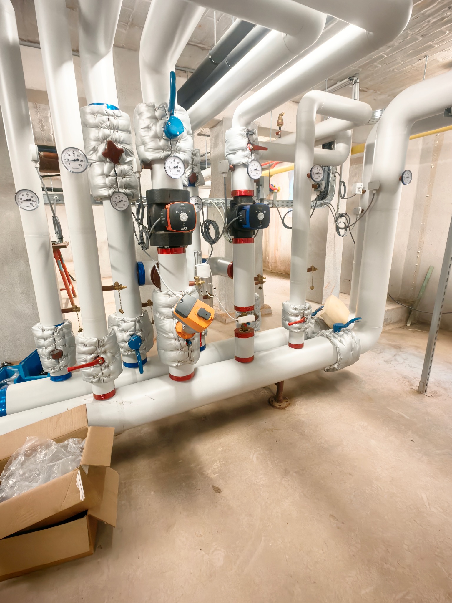 Białe rury instalacyjne z zaworami i manometrami w kotłowni, częściowo zaizolowane, z widocznym kartonem na podłodze. Ujęcie z dołu, widok na sufit.