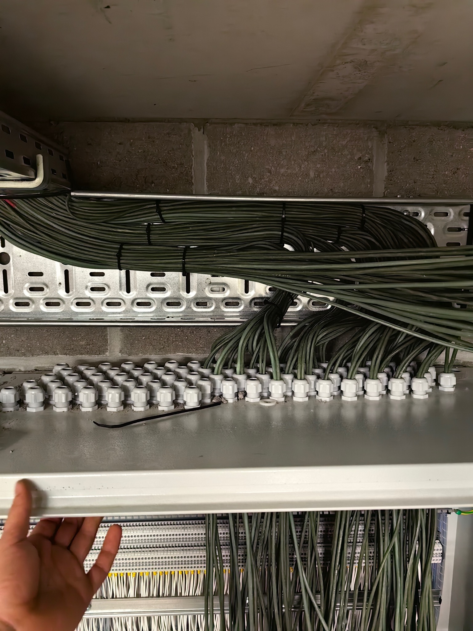 Rozbudowana instalacja elektryczna w szafie sterowniczej. Widoczne liczne przewody w peszlach, listwy zaciskowe i metalowe korytka kablowe. Ręka przytrzymuje element szafy.