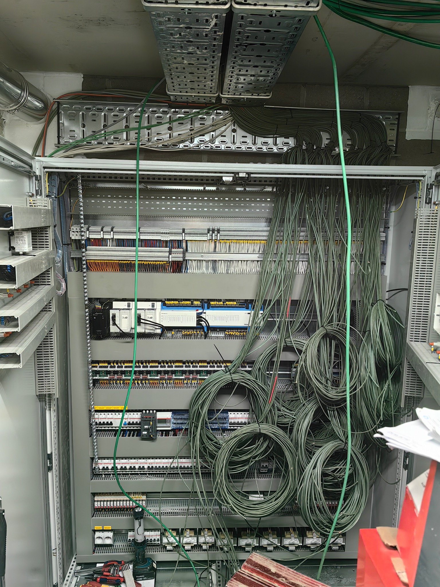 Rozdzielnica elektryczna w trakcie montażu, widoczne liczne kable, listwy zaciskowe i elementy sterujące. Kable ułożone w pętle, narzędzia na dole szafy.