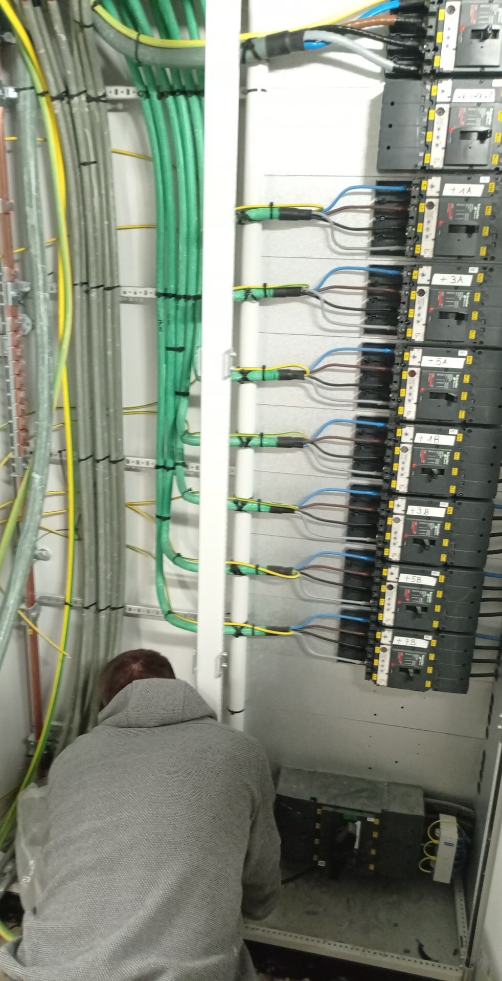 Elektryk w szarej bluzie, w trakcie montażu rozdzielnicy elektrycznej z licznymi przewodami i zabezpieczeniami. Widoczne kable w różnych kolorach i oznaczenia na urządzeniach.
