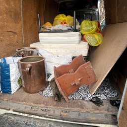 Konrad Blachaniec - Wnętrze furgonetki załadowane różnorodnymi przedmiotami: materace, worki, donica, deska, krzesła. Widoczne elementy wyposażenia pojazdu i podłoga.