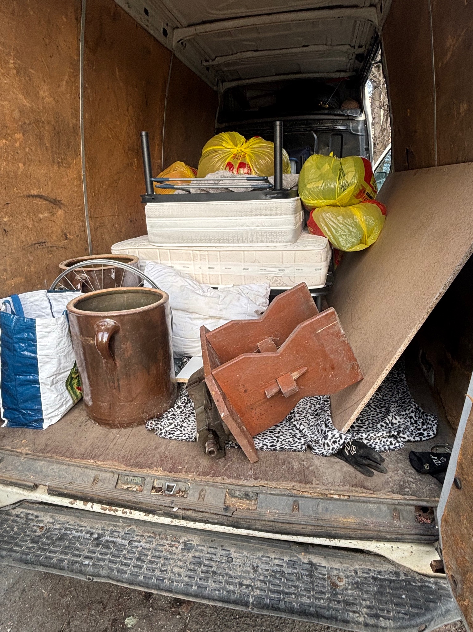 Wnętrze furgonetki załadowane różnorodnymi przedmiotami: materace, worki, donica, deska, krzesła. Widoczne elementy wyposażenia pojazdu i podłoga.