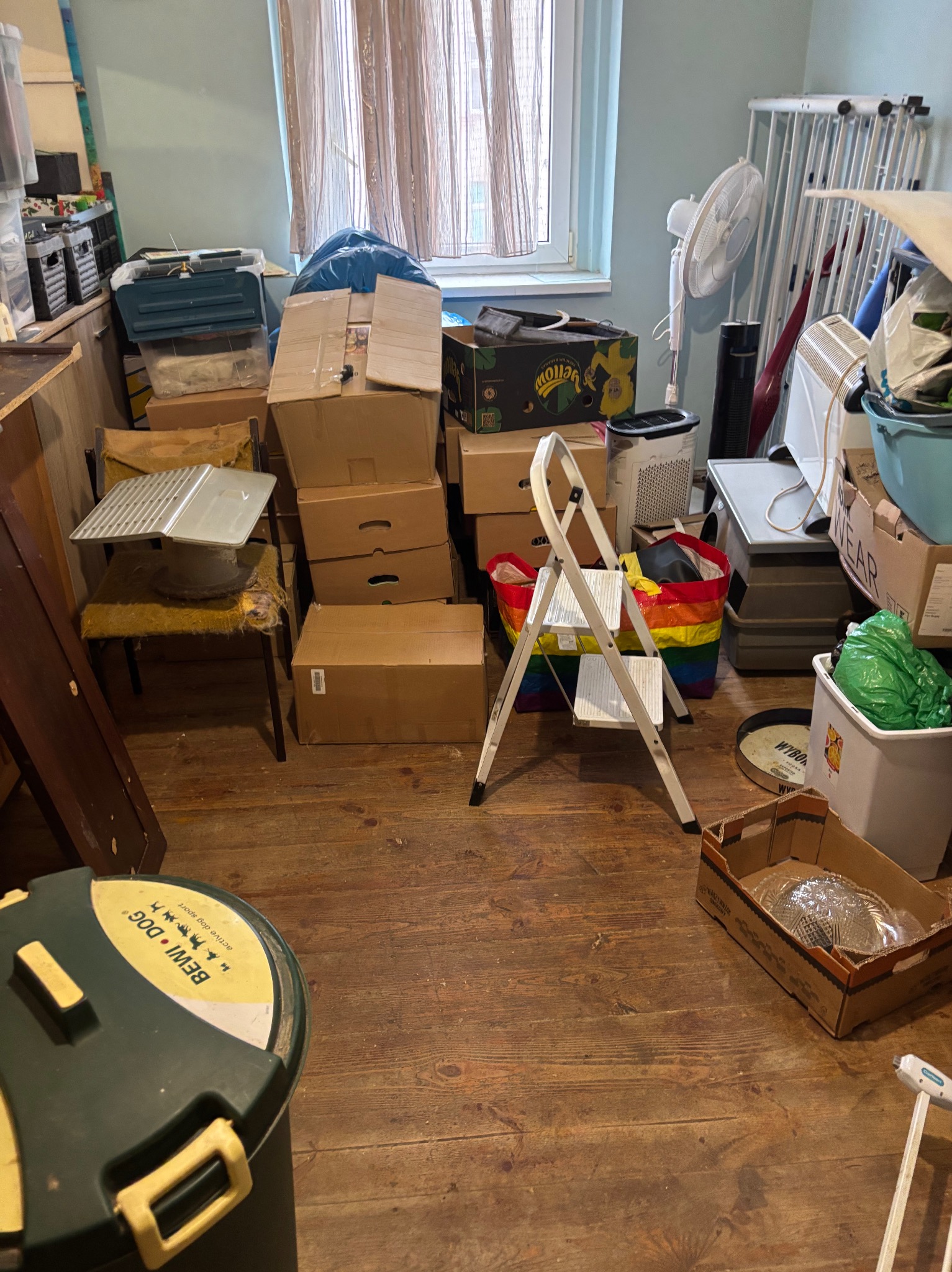 Pokój zagracony kartonami, torbami i różnymi przedmiotami przed przeprowadzką. Widoczna drabina, wentylator i drewniana podłoga.