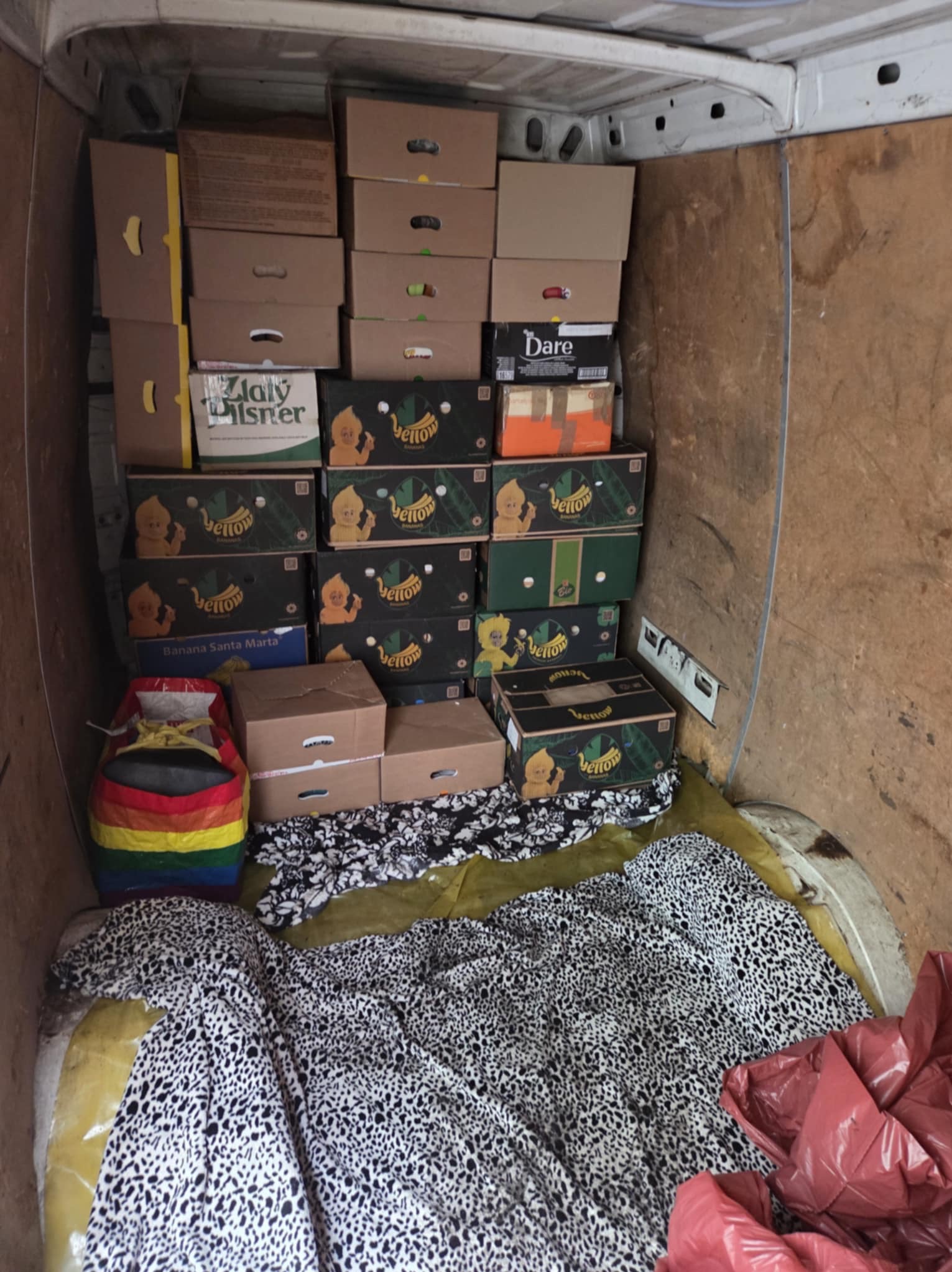 Wnętrze furgonetki wypełnione kartonami, niektóre z logo 'Yellow Banana Santa Marta', oraz przedmiotami osobistymi, w tym kolorową torbą i wzorzystą pościelą.