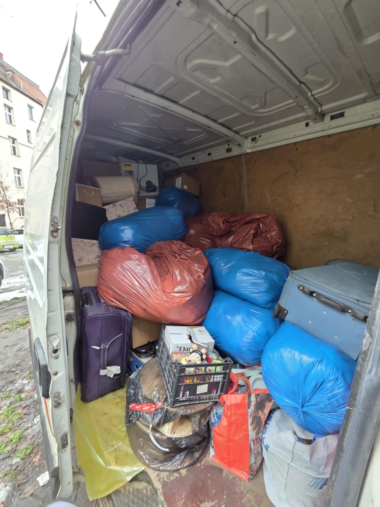 Wnętrze białego busa wypełnione po brzegi workami w kolorze niebieskim i czerwonym, kartonami i walizkami podczas przeprowadzki. Widok z zewnątrz na załadowany pojazd.