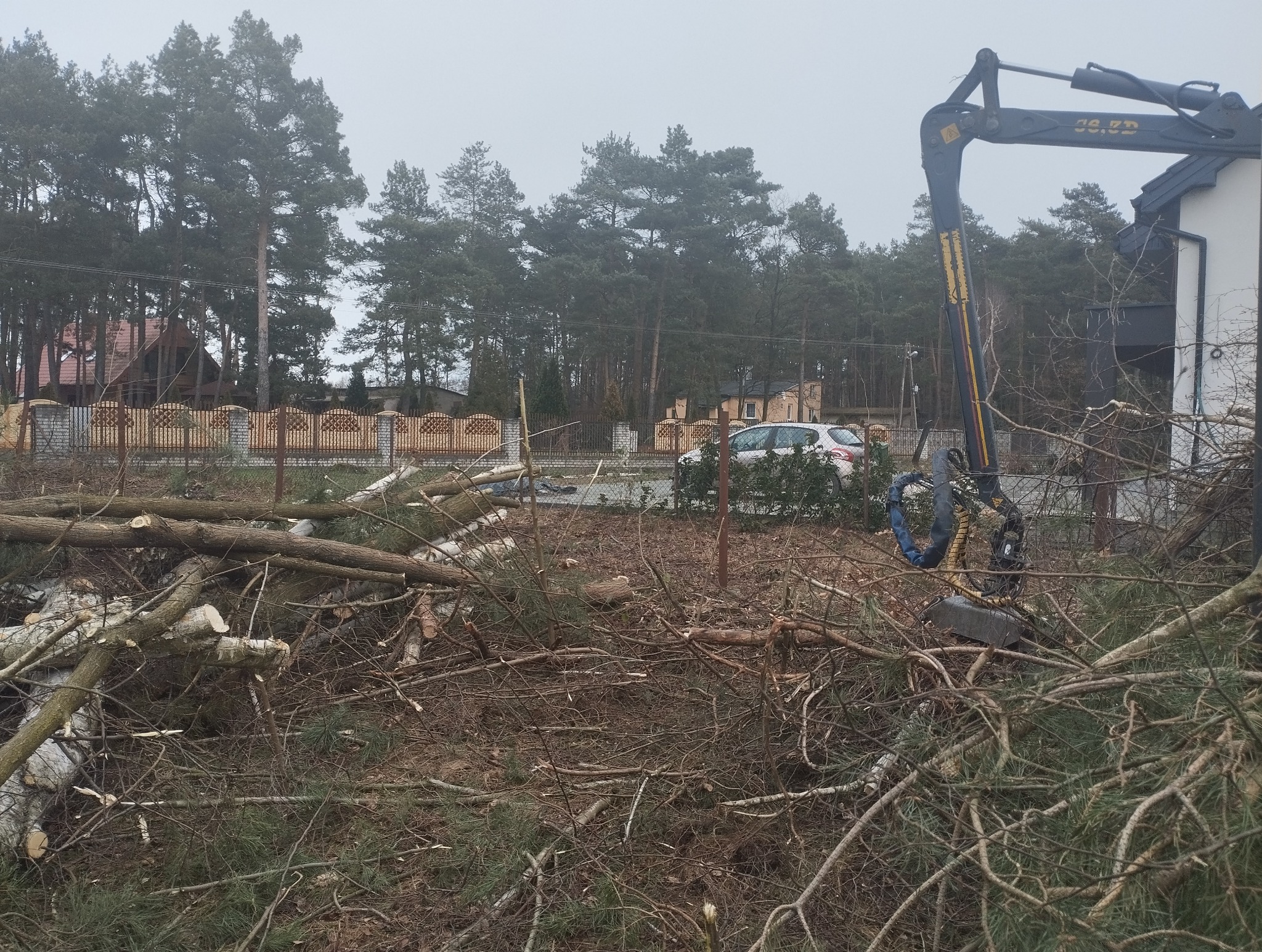 Teren po wycince drzew z porozrzucanymi gałęziami i pniem, w tle dom, ogrodzenie i samochód. Widoczny ciężki sprzęt do wycinki. Pochmurny dzień.