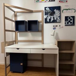 Vitek Olejnik - Białe biurko z drewnianą nadbudową i granatowymi szafkami, uzupełnione o półki i oświetlenie, na tle ściany z dekoracjami.