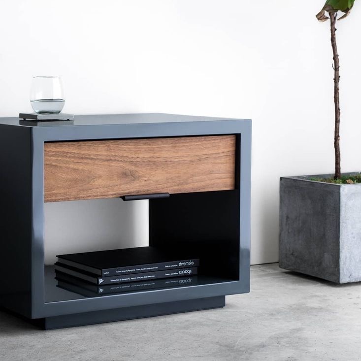 Stolik nocny z szufladą z drewna orzechowego i szarą ramą, na blacie szklanka wody, na półce książki, obok doniczka z rośliną. Minimalistyczny design.