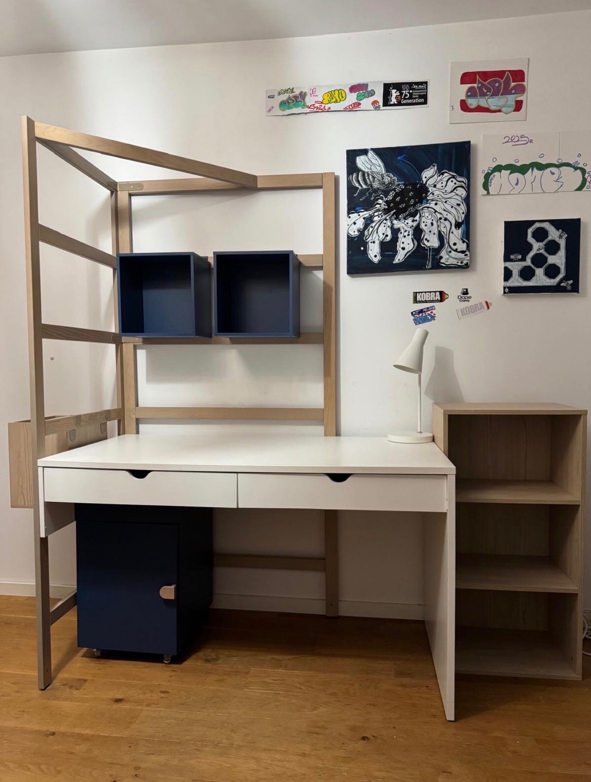Białe biurko z drewnianą nadbudową i granatowymi szafkami, uzupełnione o półki i oświetlenie, na tle ściany z dekoracjami.
