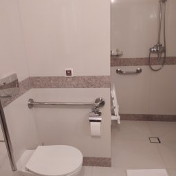 BET-MAR Beata Stępień - Jasna łazienka z toaletą, uchwytami dla niepełnosprawnych i kabiną prysznicową. Wykończenie w odcieniach bieli i beżu. Widoczny ręcznik na podłodze.