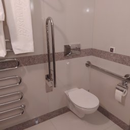 BET-MAR Beata Stępień - Nowoczesna, jasna łazienka z wiszącą toaletą, uchwytami dla niepełnosprawnych i grzejnikiem drabinkowym. Minimalistyczny design z akcentami w kolorze beżowym.