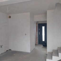 BET-MAR Beata Stępień - Surowe wnętrze w trakcie prac wykończeniowych: szpachlowane ściany, betonowa podłoga, widoczne instalacje elektryczne i fragment schodów. Drzwi wejściowe w tle.