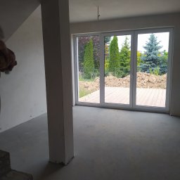 BET-MAR Beata Stępień - Surowe wnętrze z dużym oknem tarasowym i widokiem na ogród. Ściany przygotowane do malowania, widoczny betonowy filar i surowa podłoga. Etap prac wykończeniowych.