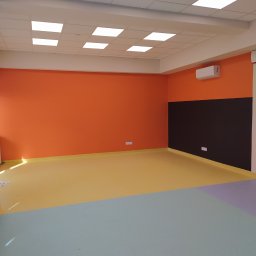 BET-MAR Beata Stępień - Wnętrze po malowaniu: pomarańczowe ściany, czarny pas, wielokolorowa podłoga (żółty, szary, fioletowy). Sufit z kasetonami i klimatyzacja. Widok na odnowione pomieszczenie.