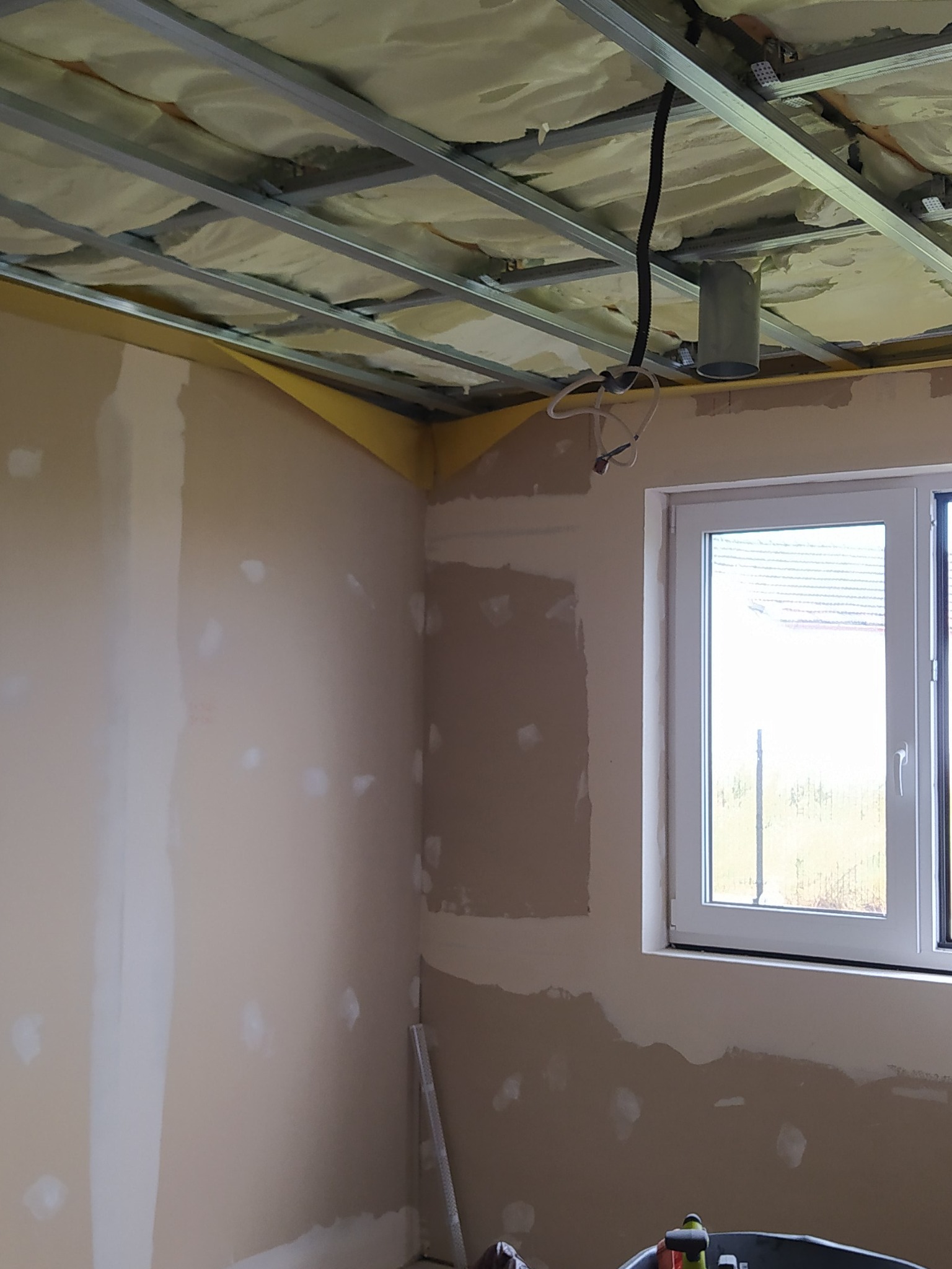 Wnętrze w trakcie prac wykończeniowych: szpachlowane ściany, sufit podwieszany z izolacją, okno. Widoczne narzędzia w wiadrze. Etap przygotowania do malowania.