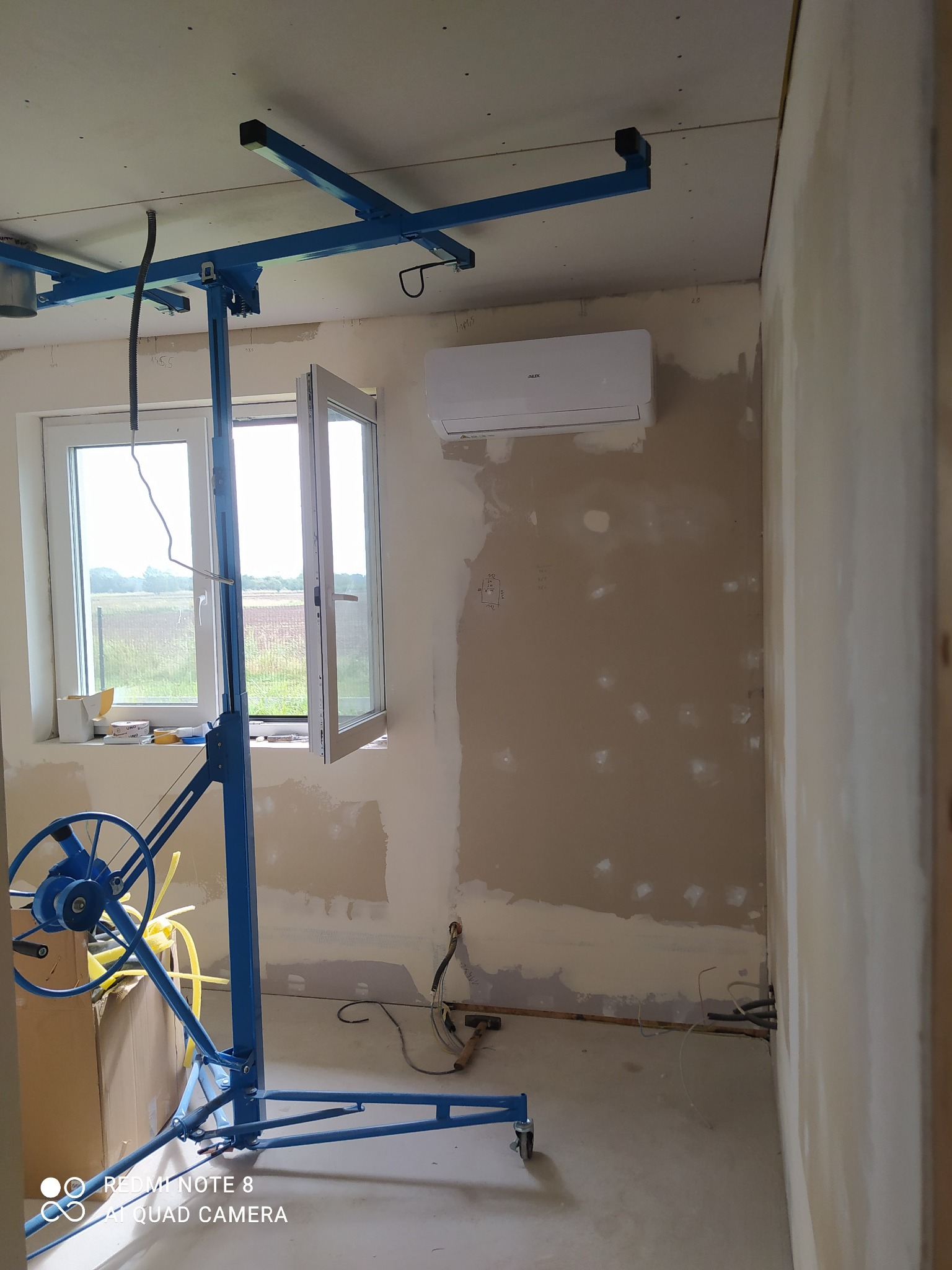 Wnętrze w trakcie prac wykończeniowych: ściany po szpachlowaniu, sufit z płyt karton-gips, podnośnik do płyt, okno z widokiem na zewnątrz, klimatyzator na ścianie.