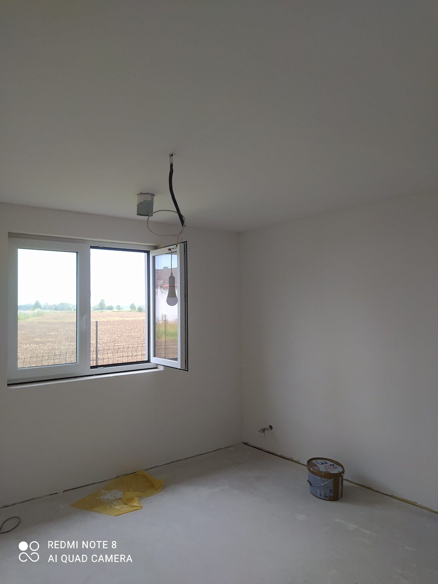 Pusty pokój po szpachlowaniu, przygotowany do malowania. Widoczne gładkie, białe ściany, otwarte okno z widokiem na zewnątrz i wiadro z farbą w rogu.