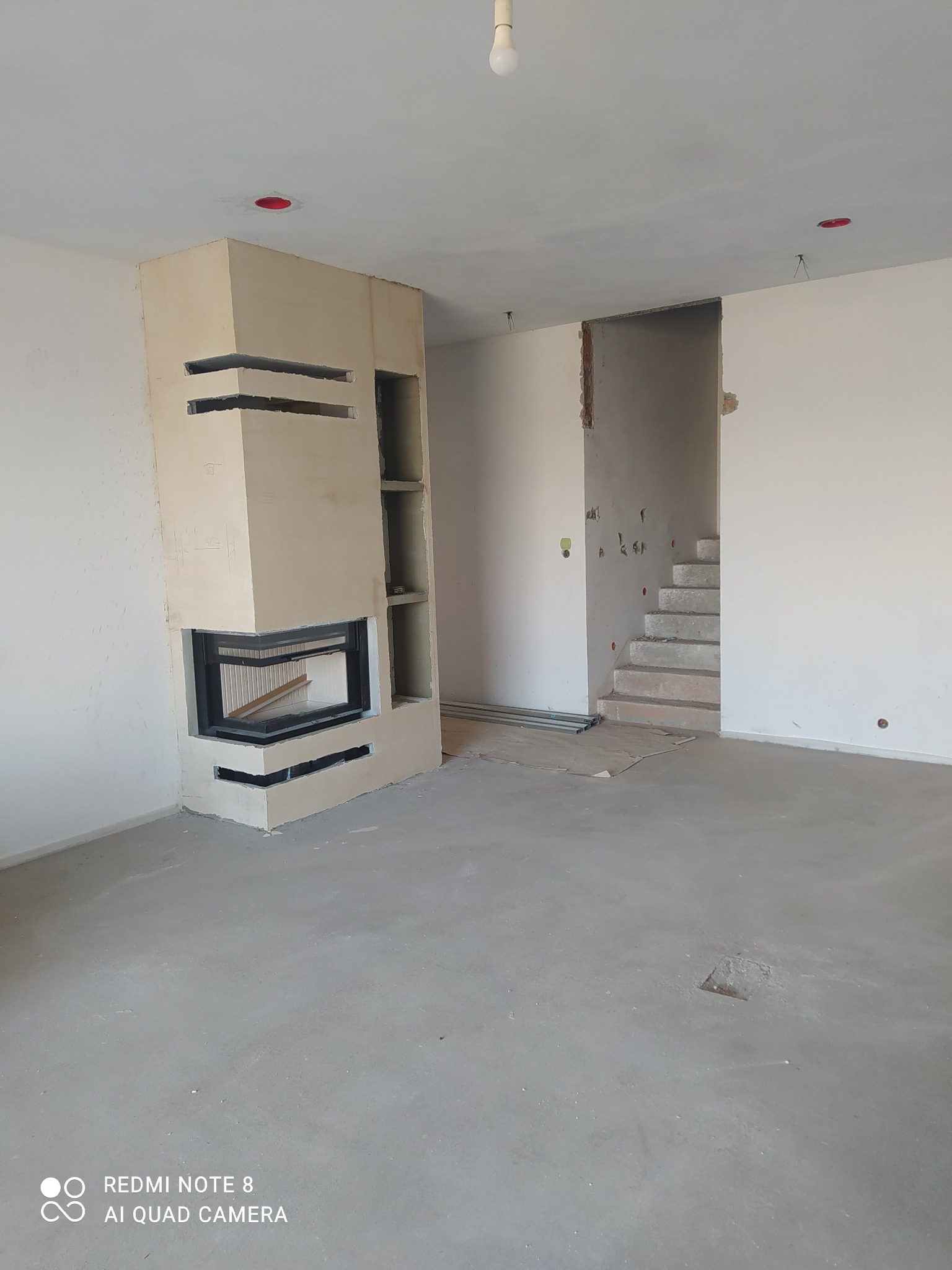 Surowy stan wnętrza z kominkiem w zabudowie, widoczne schody w tle i instalacje elektryczne w suficie. Ściany przygotowane do malowania, podłoga wylewka.