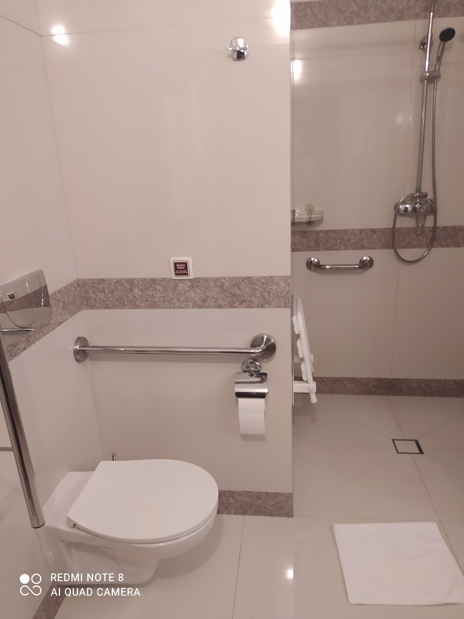 Jasna łazienka z toaletą, uchwytami dla niepełnosprawnych i kabiną prysznicową. Wykończenie w odcieniach bieli i beżu. Widoczny ręcznik na podłodze.