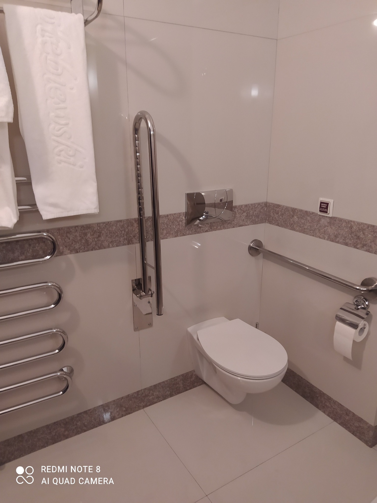 Nowoczesna, jasna łazienka z wiszącą toaletą, uchwytami dla niepełnosprawnych i grzejnikiem drabinkowym. Minimalistyczny design z akcentami w kolorze beżowym.