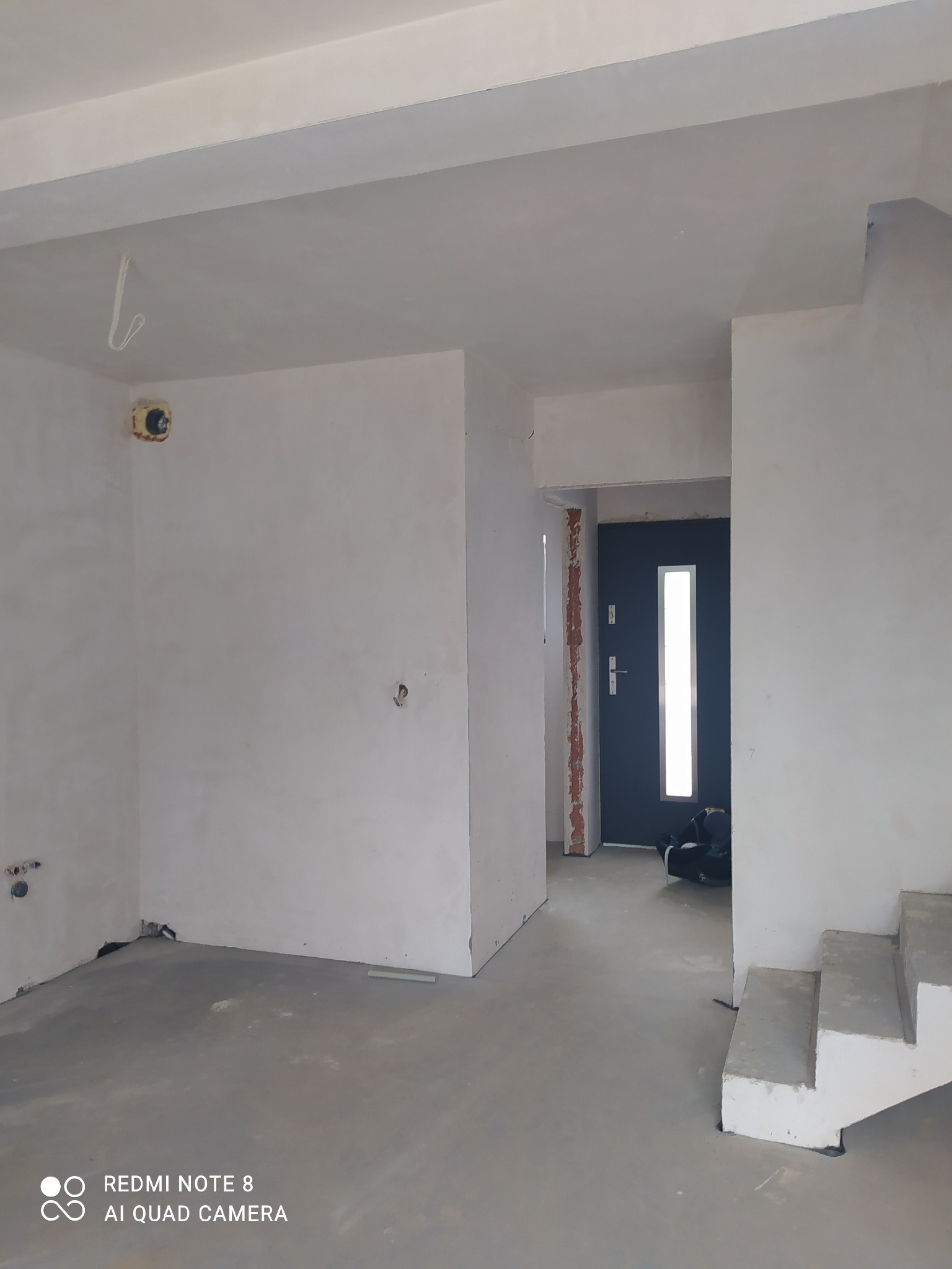 Surowe wnętrze w trakcie prac wykończeniowych: szpachlowane ściany, betonowa podłoga, widoczne instalacje elektryczne i fragment schodów. Drzwi wejściowe w tle.