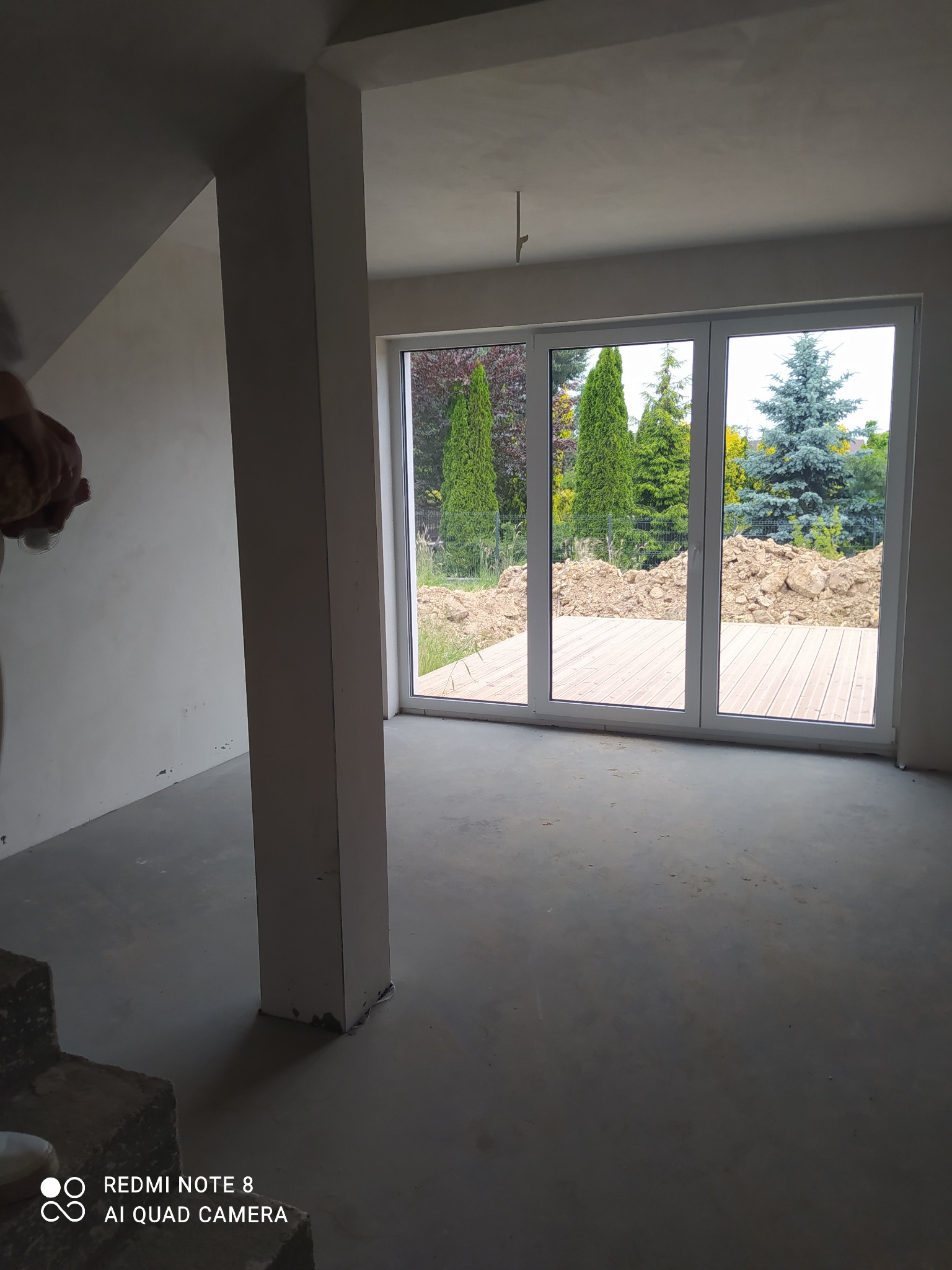 Surowe wnętrze z dużym oknem tarasowym i widokiem na ogród. Ściany przygotowane do malowania, widoczny betonowy filar i surowa podłoga. Etap prac wykończeniowych.