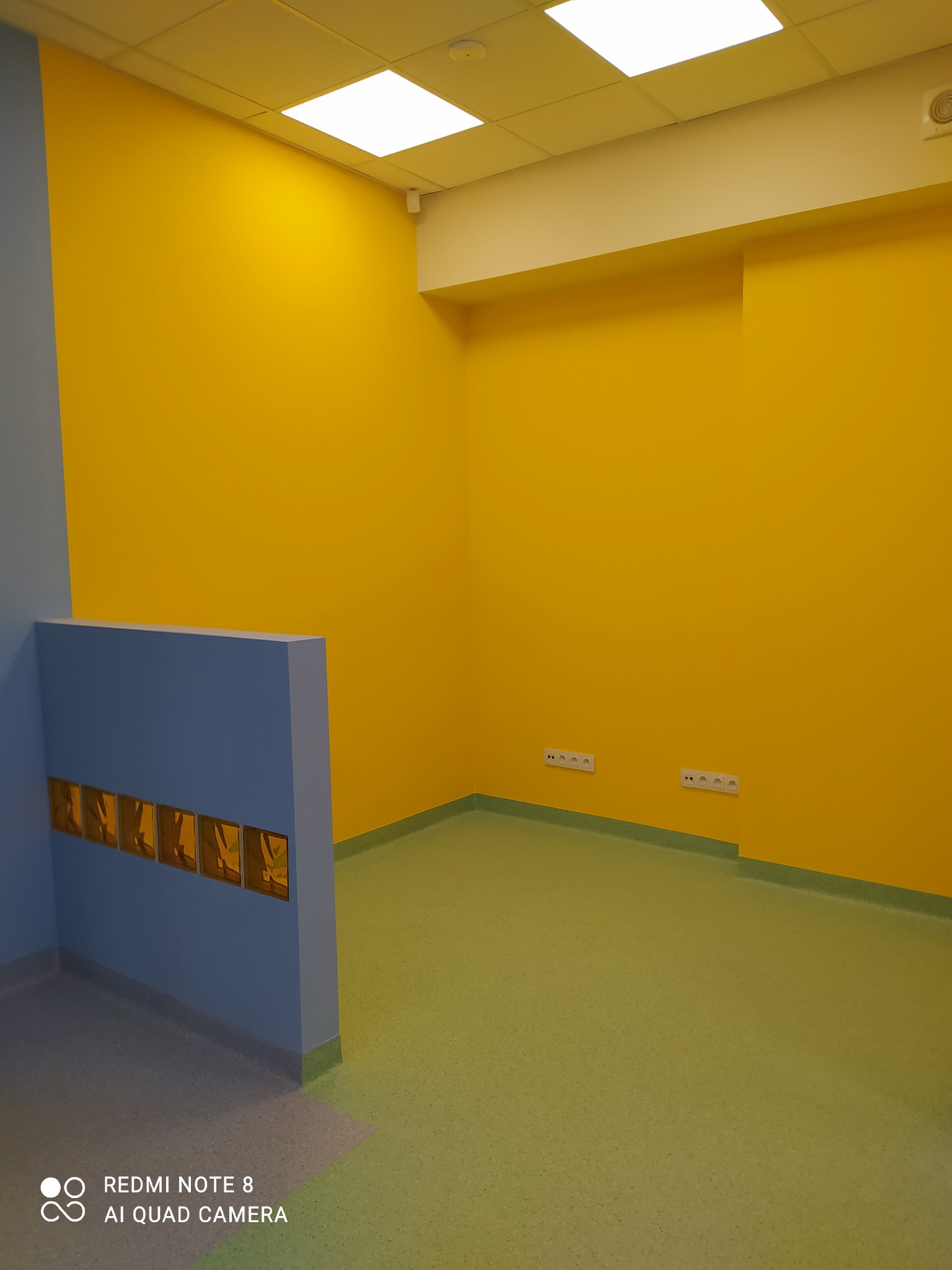Pomalowane na żółto ściany w pomieszczeniu z zieloną podłogą i niebieskim elementem architektonicznym z wieszakami. Widoczne gniazdka elektryczne.