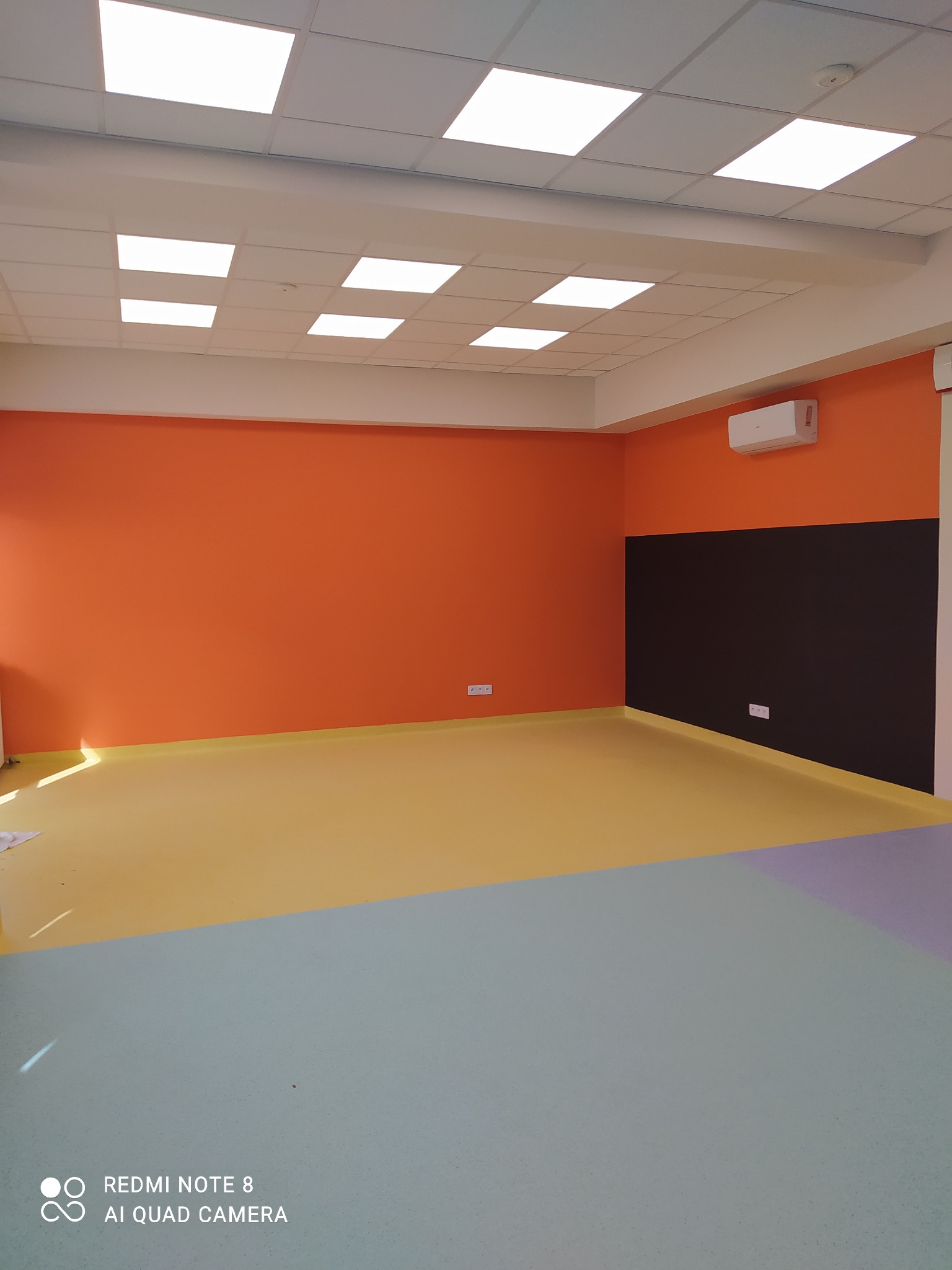 Wnętrze po malowaniu: pomarańczowe ściany, czarny pas, wielokolorowa podłoga (żółty, szary, fioletowy). Sufit z kasetonami i klimatyzacja. Widok na odnowione pomieszczenie.