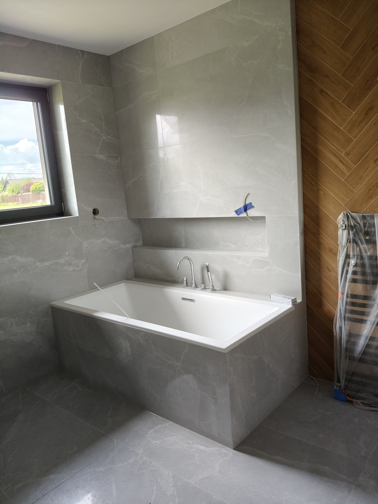 Nowoczesna łazienka z wanną obłożoną szarym marmurem, wnęka w ścianie, drewniana ściana w jodełkę, grzejnik owinięty folią. Widok na zewnątrz przez okno.