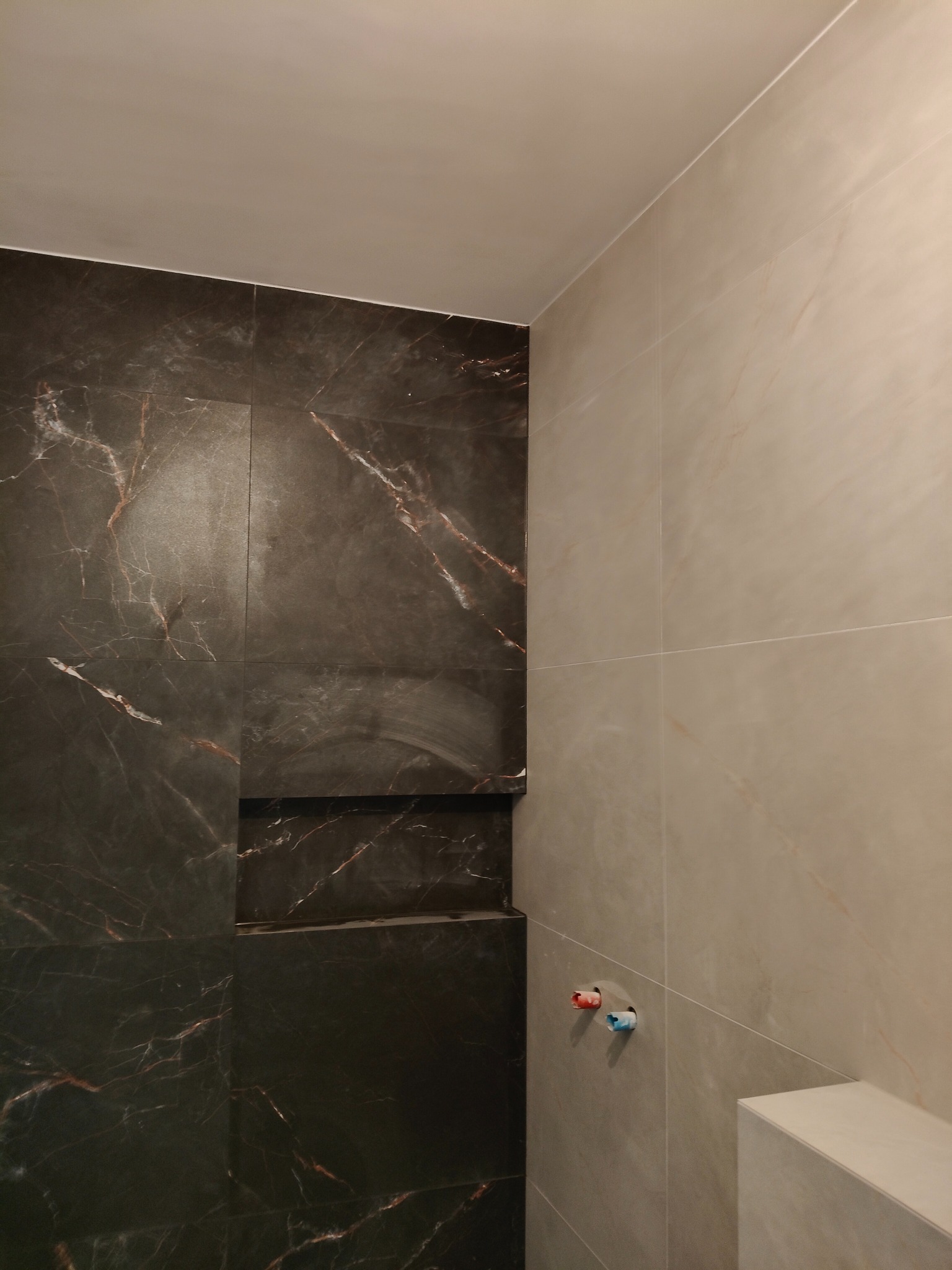 Wykończona wnęka prysznicowa z ciemnymi płytkami imitującymi marmur i jasnymi płytkami na ścianie. Widoczne wystające rury do baterii.