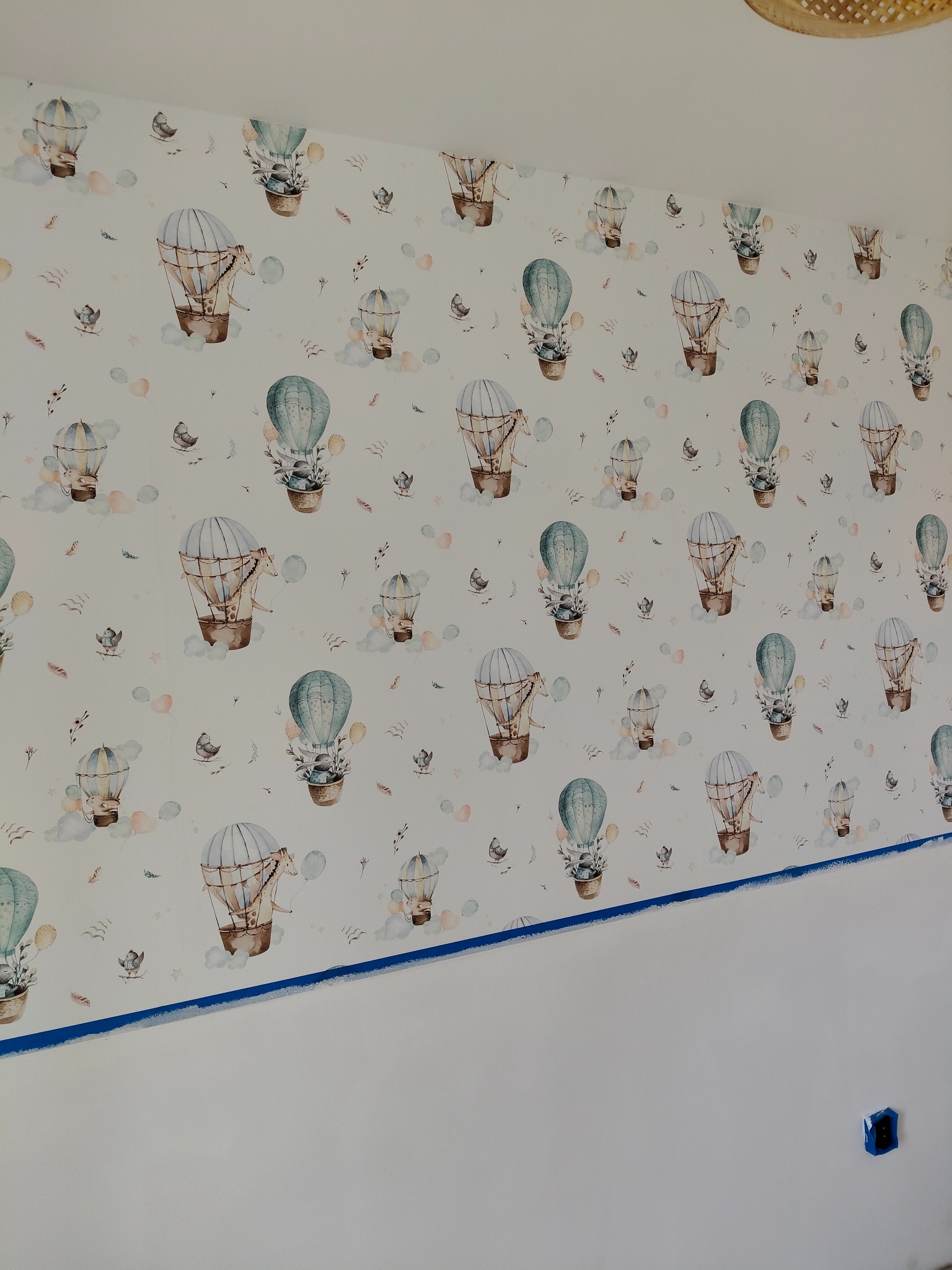 Ściana z tapetą w dziecięcym pokoju w Krakowie, wzór w balony i zwierzęta, zabezpieczona niebieską taśmą malarską przy suficie i podłodze. Widoczny fragment gniazdka elektrycznego.