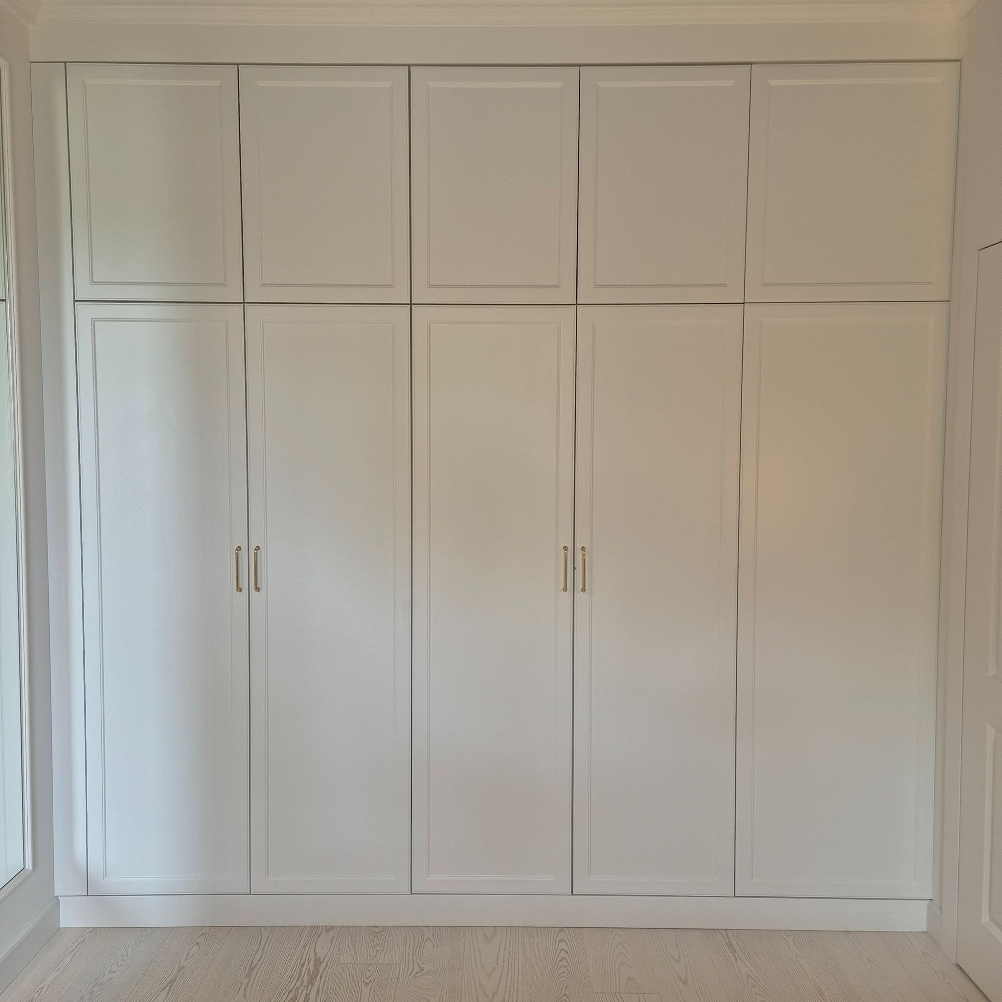 Biała, zabudowana szafa wnękowa z frezowanymi frontami i złotymi uchwytami, dopasowana do listew przypodłogowych i sufitowych. Jasna podłoga z paneli.
