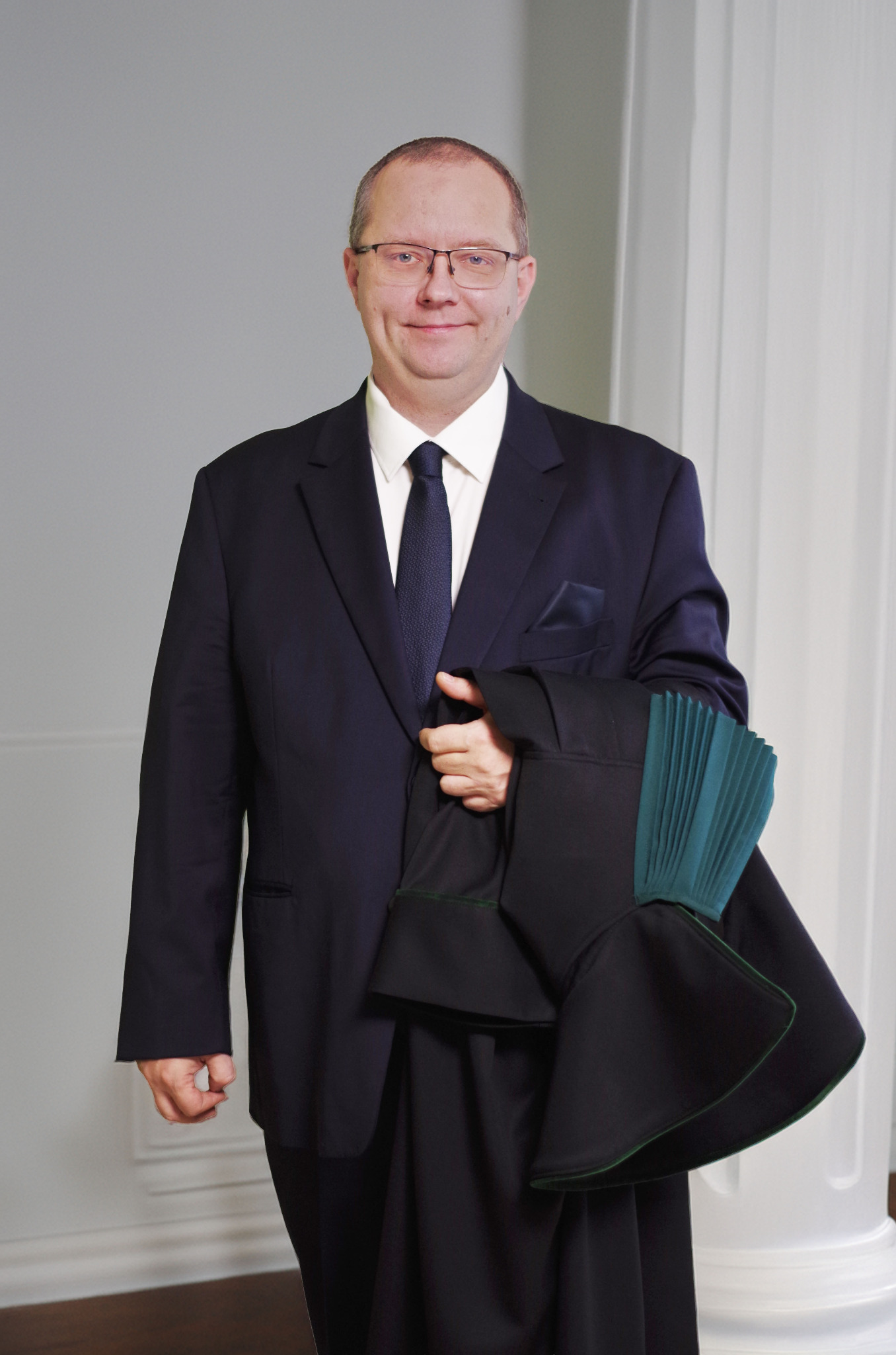 Portret adwokata w todze, trzymającego ją w ręku, na tle kolumny. Mężczyzna w garniturze i okularach, uśmiechnięty, patrzy wprost. Profesjonalny wizerunek prawnika.