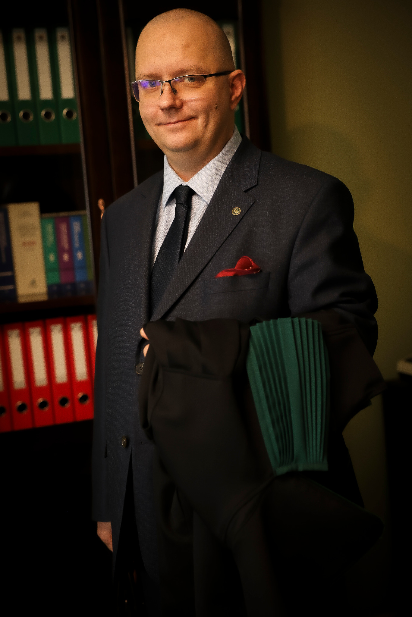 Uśmiechnięty adwokat w garniturze, trzymający togę na tle regału z segregatorami. Widoczna odznaka adwokacka i poszetka w butonierce.