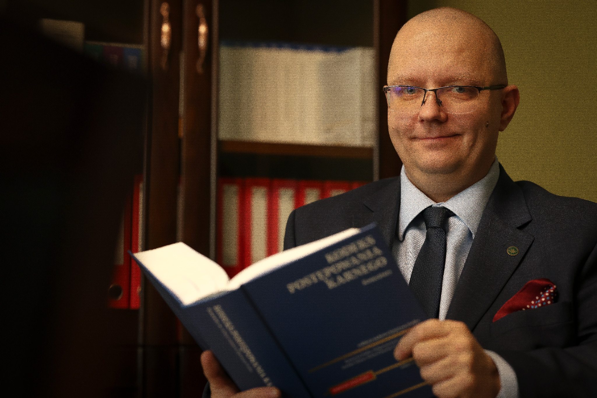 Portret adwokata w okularach, trzymającego otwarty kodeks karny na tle regału z aktami. Widoczny garnitur i poszetka w kieszonce.