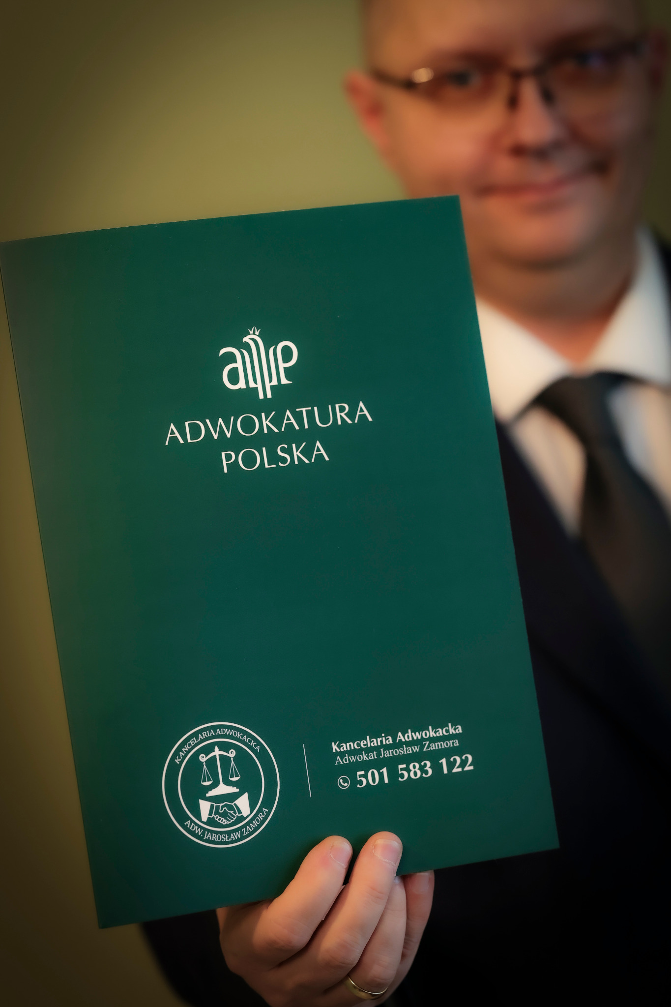 Zielony folder z logo Adwokatury Polskiej trzymany przez mężczyznę w okularach i garniturze. Widoczny numer telefonu do Kancelarii Adwokackiej Jarosław Zamora.
