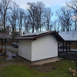 KoMa Energy Sp. z o.o. - Niedokończony budynek z białych bloczków z panelami słonecznymi na dachu, otoczony drzewami i zielenią. Widoczne samochody i osoba w odblaskowej kurtce.