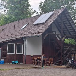 KoMa Energy Sp. z o.o. - Dom z brązowym dachem i panelami słonecznymi, otoczony zielenią. Widoczne rowery i osoba siedząca przy drewnianym stole. Ujęcie z zewnątrz, w dzień.