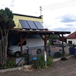 KoMa Energy Sp. z o.o. - Instalacja solarna na drewnianej wiacie garażowej przy żółtym budynku. Biały van zaparkowany wewnątrz, widoczne ogrodzenie i samochody w tle.