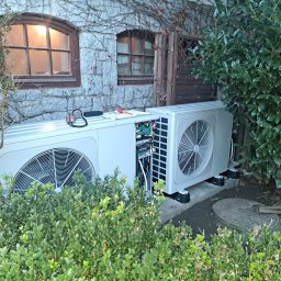 KoMa Energy Sp. z o.o. - Dwie jednostki zewnętrzne pompy ciepła, jedna z otwartą obudową, widoczne okablowanie, na tle kamiennej ściany budynku z oknami i zielenią.