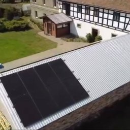 KoMa Energy Sp. z o.o. - Panele słoneczne na dachu budynku w Legnicy, widok z góry. Cztery czarne panele na srebrnym, blaszanym dachu. W tle budynek z muru pruskiego i zielony trawnik.