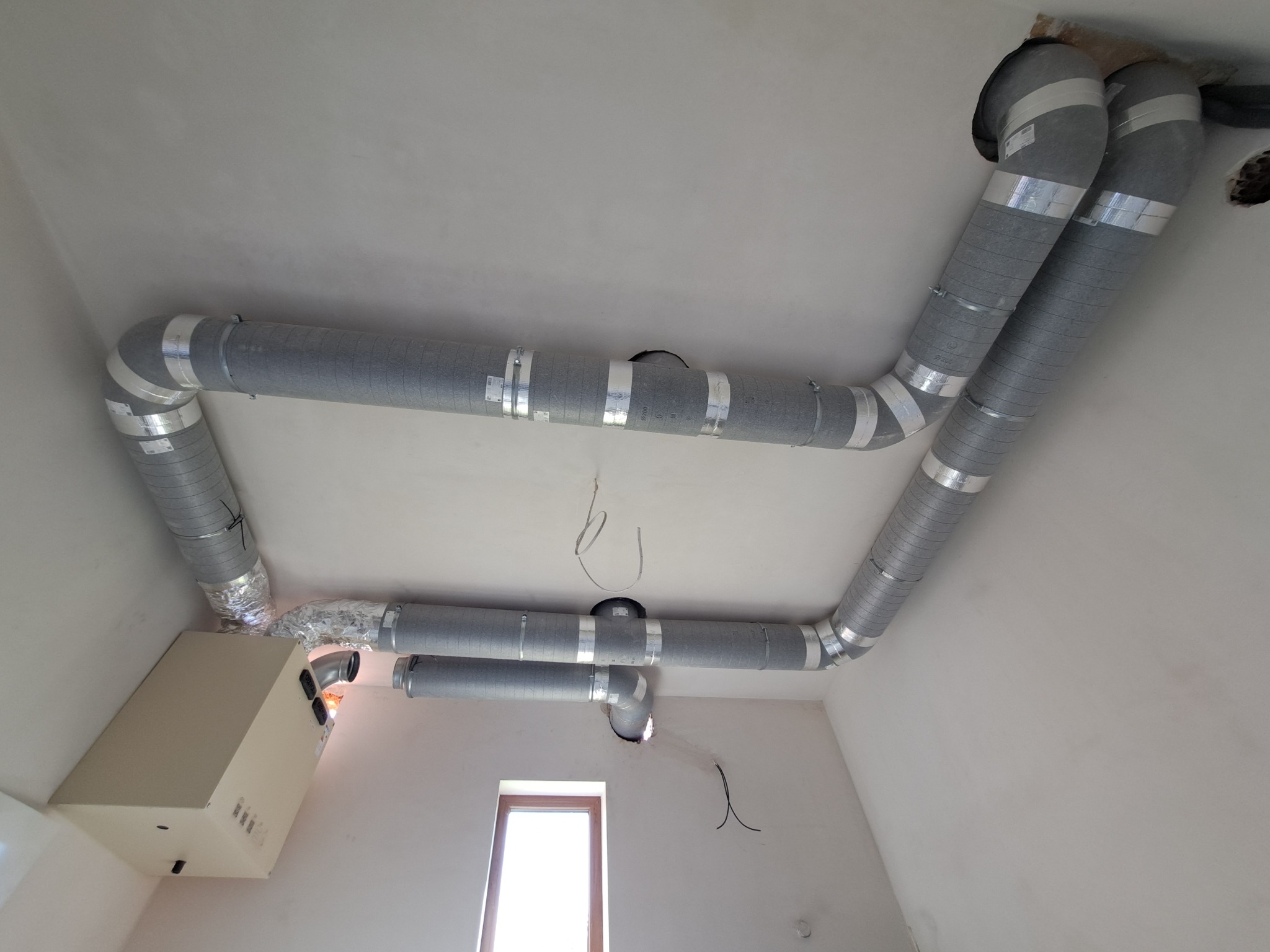 Szare, elastyczne rury wentylacyjne z taśmą mocującą, zamontowane pod sufitem w jasnym pomieszczeniu z oknem, część instalacji rekuperacji.