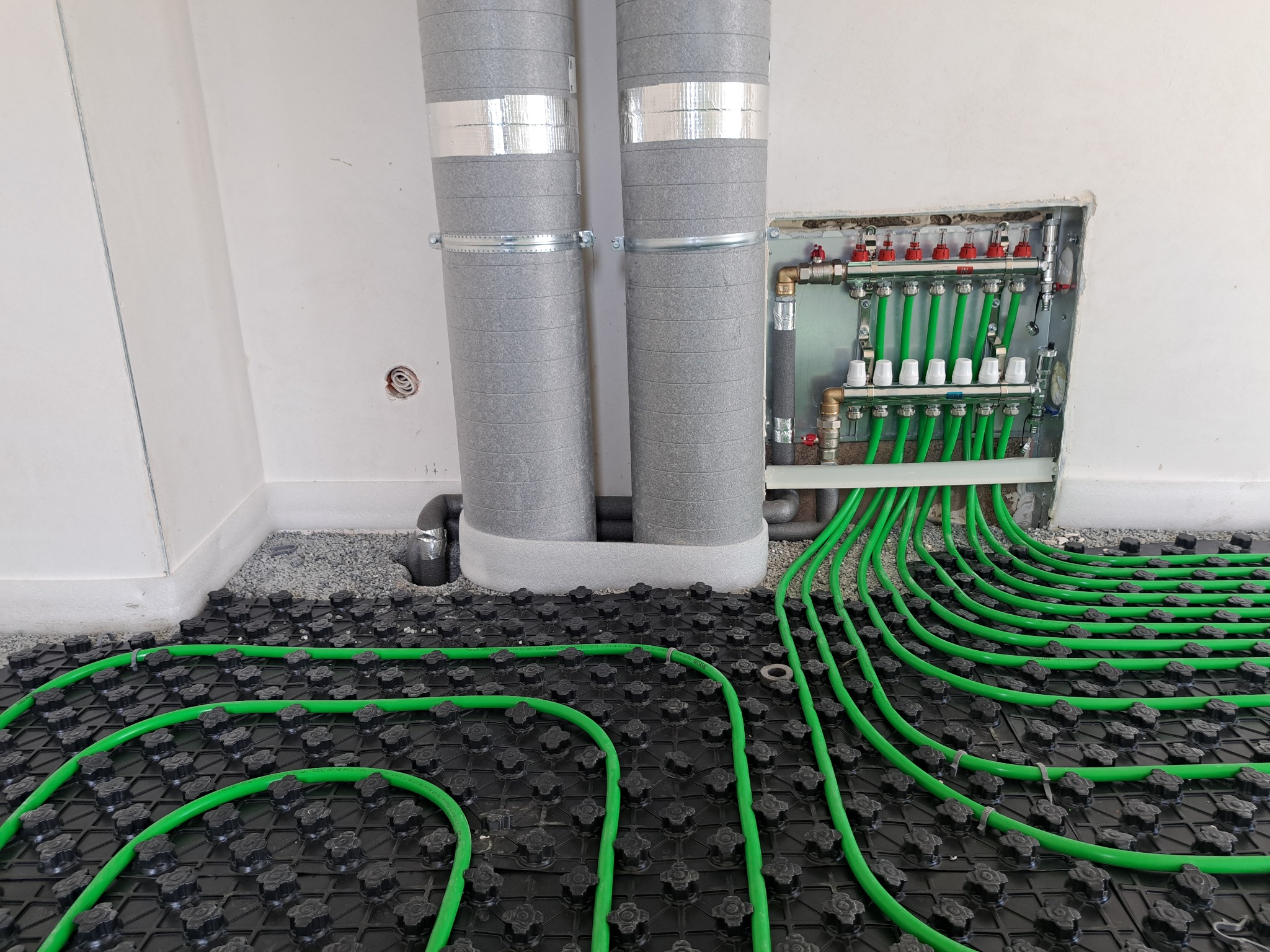 Zielone rury ogrzewania podłogowego ułożone na czarnej macie z rozdzielaczem w ścianie i izolowanymi pionami. Widok z góry, instalacja w trakcie realizacji.