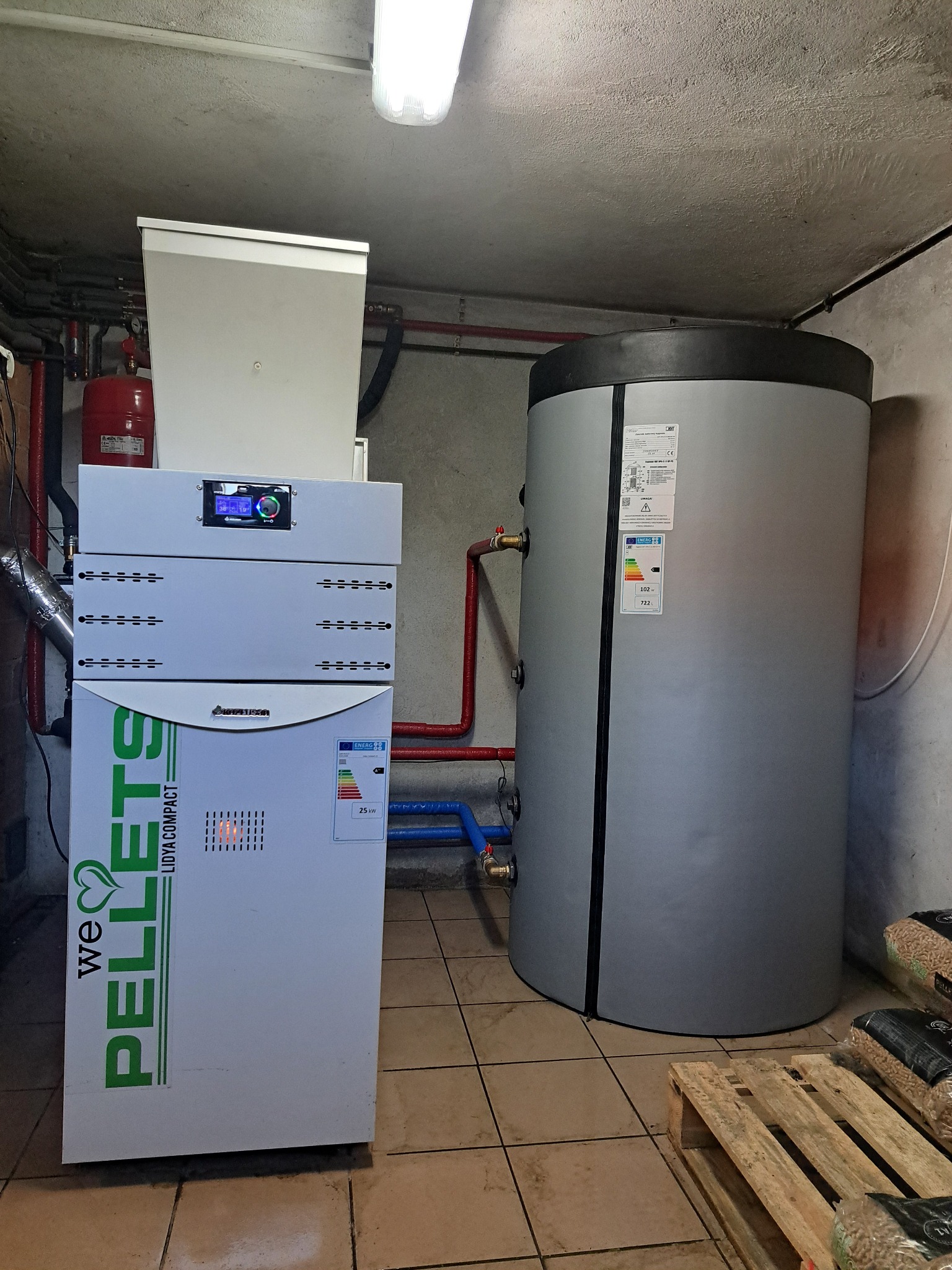Kotłownia z kotłem na pellet i zasobnikiem ciepłej wody, widoczne worki z pelletem. Instalacja w pomieszczeniu z płytkami na podłodze i oświetleniem sufitowym.